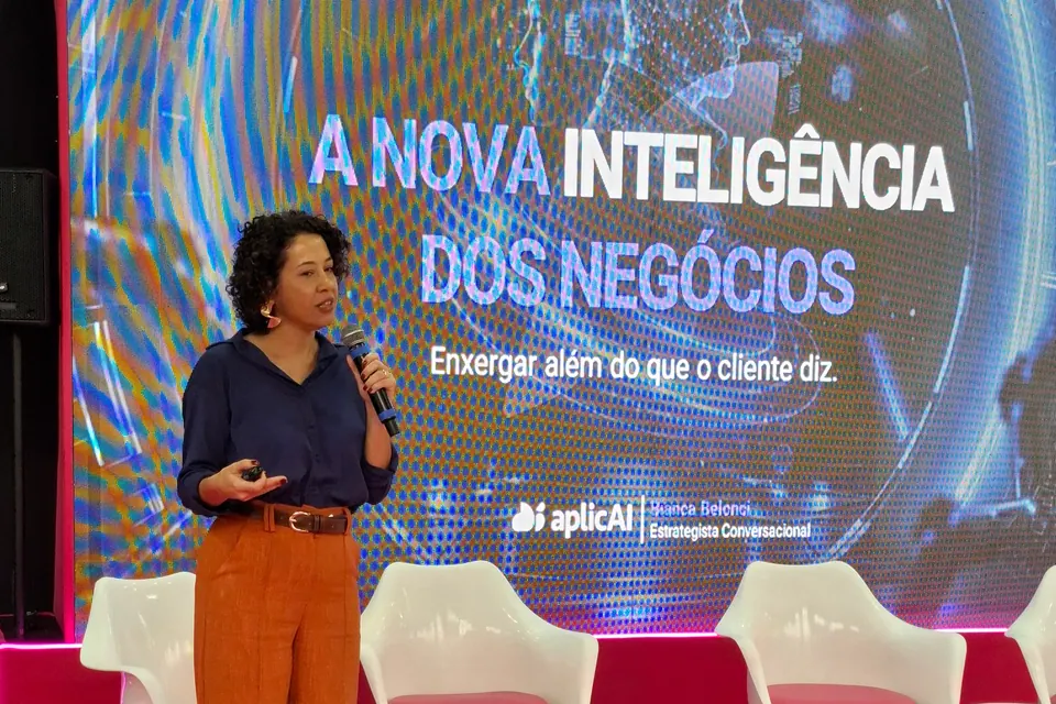 Pessoa de blusa azul e calça laranja com microfone diante de uma tela no palco.