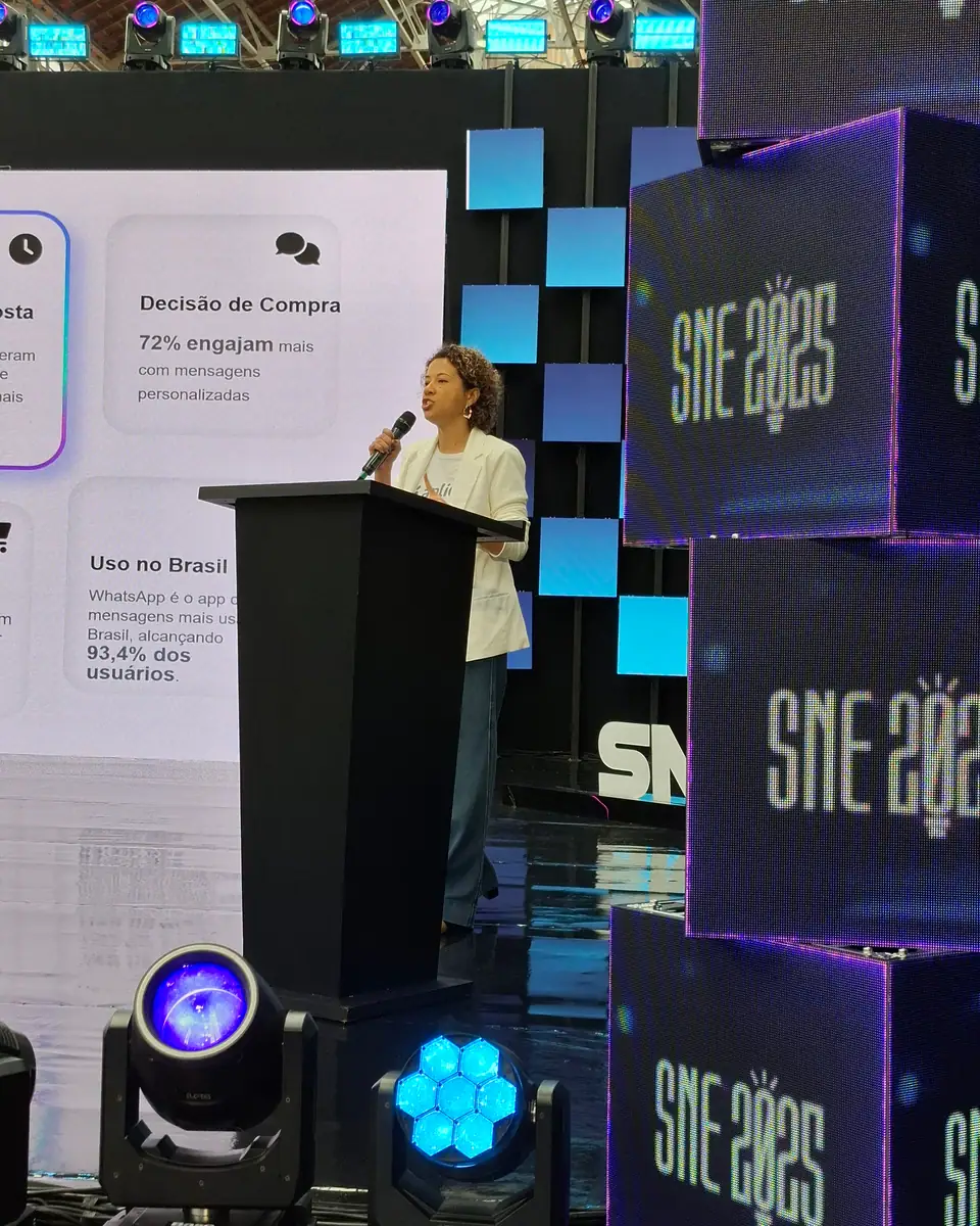 Pessoa de blazer claro falando ao microfone em uma tribuna, com a identificação SNE 2025 ao fundo.
