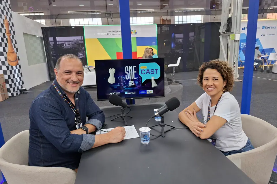 Duas pessoas sentadas à mesa com microfones e uma tela com a identificação SNE Cast.